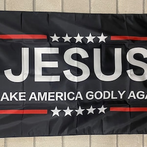 May include: Black flag with the text "JESUS MAKE AMERICA GODLY AGAIN" in white letters. There are five white stars above and below the text.
