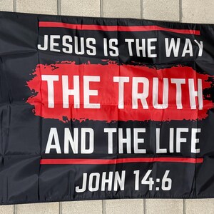 May include: Black flag with white text that reads "JESUS IS THE WAY THE TRUTH AND THE LIFE JOHN 14:6" in a bold font. The words "THE TRUTH" are in a red rectangular block. The flag has a white border and grommets.