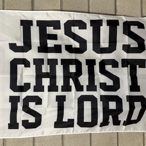 May include: White flag with the text "JESUS CHRIST IS LORD" in bold, black, block letters. The flag has a grommet and a white cord for hanging. The flag is rectangular and appears to be made of a lightweight fabric.