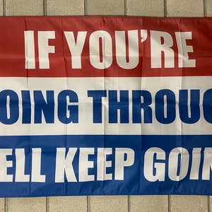 Puede incluir: Una bandera rectangular con una combinación de colores rojo, blanco y azul. La bandera presenta el texto "IF YOU'RE GOING THROUGH HELL KEEP GOING" en letras blancas en negrita. La bandera cuelga contra una pared texturizada de color claro.