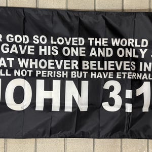 May include: Black flag with white text. The text reads: "FOR GOD SO LOVED THE WORLD THAT HE GAVE HIS ONE AND ONLY SON, THAT WHOEVER BELIEVES IN HIM SHALL NOT PERISH BUT HAVE ETERNAL LIFE. JOHN 3:16."