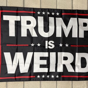 May include: Black flag with the words "TRUMP IS WEIRD" in large white capital letters. The text is framed by a red border and accented with white stars. The flag has grommets for hanging.