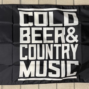 Puede incluir: Bandera negra con texto blanco "COLD BEER & COUNTRY MUSIC". La bandera tiene un borde blanco en el lado izquierdo y está sobre una superficie de baldosas beige. La bandera rectangular tiene una arandela en la esquina superior izquierda.