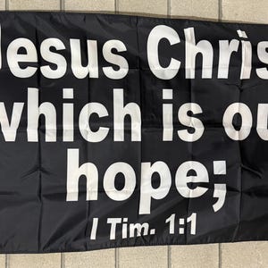 May include: Black flag with white text that reads "Jesus Christ, which is our hope; I Tim. 1:1". The flag has a white border and grommets for hanging. The flag is rectangular and made of a lightweight material.
