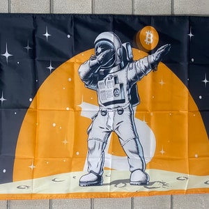 Könnte beinhalten: Eine schwarz-orangefarbene Flagge mit einem Cartoon-Astronauten, der den Dab macht. Der Astronaut steht auf einer mondähnlichen Oberfläche mit einem großen gelben Mond im Hintergrund. Der Astronaut trägt einen weißen Raumanzug und einen Helm. Der Mond hat ein Bitcoin-Symbol darauf.
