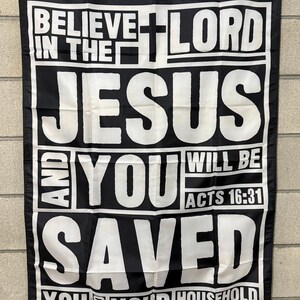 May include: Black flag with white text that reads "BELIEVE IN THE LORD JESUS AND YOU WILL BE SAVED YOU AND YOUR HOUSEHOLD" with a cross graphic. The text "ACTS 16:31" is also included. The flag has a white border and gold grommets.