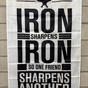 May include: White flag with black text that reads "IRON SHARPENS IRON SO ONE FRIEND SHARPENS ANOTHER" with a blacksmith anvil graphic at the top. The text "Proverbs 27:17" is at the bottom.