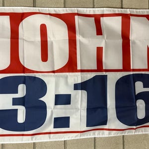 May include: White flag with red and blue text. The word "JOHN" is in large white letters on a red background. Below, the numbers "3:16" are in blue on a white background, framed by a red border.
