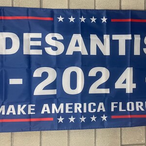 May include: A blue political campaign flag with white text that reads "DESANTIS - 2024 - MAKE AMERICA FLORIDA" with red and white stars at the top and bottom.