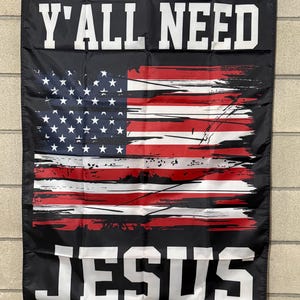 May include: Black flag with the American flag design and the words "Y'ALL NEED JESUS" in white block letters. The flag has grommets for hanging.