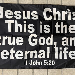 May include: Black flag with white text that reads "Jesus Christ, This is the true God, and eternal life. I John 5:20." The flag has a white border and grommets for hanging. The flag is displayed on a light-colored tiled surface.