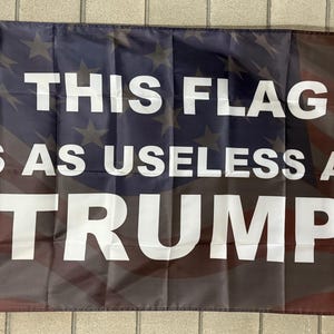 May include: A rectangular flag with a design reminiscent of the American flag. The flag features the text "THIS FLAG IS AS USELESS AS TRUMP" in large white capital letters. The background has a faded red, white, and blue color scheme.