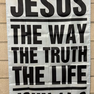 May include: White banner with black text that reads "JESUS THE WAY THE TRUTH THE LIFE JOHN 14:6". The banner is hanging against a light-colored brick wall. The text is in a bold, sans-serif font.
