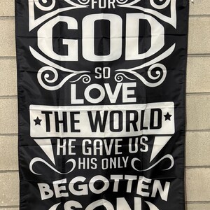 May include: Black flag with white text. The flag reads "FOR GOD SO LOVE THE WORLD HE GAVE US HIS ONLY BEGOTTEN SON JOHN 3:16". The flag has a white border and grommets at the top for hanging.