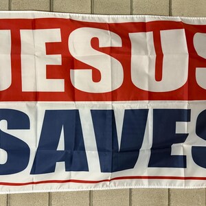 May include: A rectangular flag with the words "JESUS SAVES" in large, bold, white letters. The top section is red, and the bottom section is blue. The flag has a white border and grommets.
