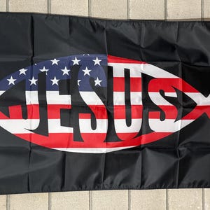 Op de afbeelding: Zwarte vlag met een vissymbool met de sterren en strepen van de Amerikaanse vlag, met het woord "JESUS" in rood en wit. De vlag heeft een witte rand aan de linkerkant.