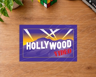 May include: A rectangular sign featuring a purple border and a graphic of purple mountains against a yellow and orange sky with white spotlights. The text HOLLYWOOD is printed in large white letters, with the word VIDEO in red below it.