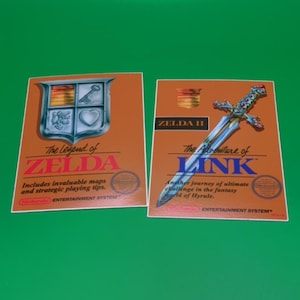 May include: Two retro video game box art prints. One features a shield with a key and heart, the text "The Legend of Zelda", and "Nintendo Entertainment System". The other shows a sword and the text "Zelda II The Adventure of Link".