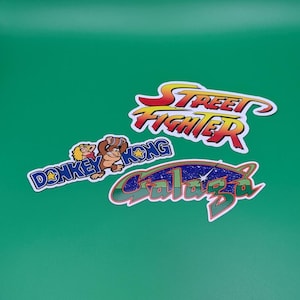 May include: Three colorful stickers featuring classic video game titles. The stickers include "Street Fighter" in red and yellow, "Donkey Kong" with a cartoon ape, and "Galaga" with a space theme, all on a green background.
