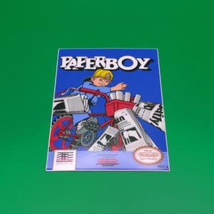 May include: A retro video game box for "Paperboy" on a green background. The cover art features a boy on a red bicycle throwing newspapers. The title "PAPERBOY" is in large white letters above the illustration. The Nintendo logo is visible.