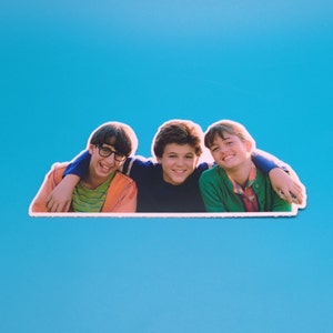 May include: Three smiling friends, two boys and a girl, are posing for a photo. The boy on the left is wearing glasses and a striped shirt. The boy in the middle is wearing a blue shirt. The girl on the right is wearing a green jacket and a patterned shirt.