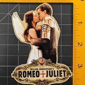 May include: A gold-coloured sticker with a decorative frame and the text "WILLIAM SHAKESPEARE'S ROMEO + JULIET". The sticker features a couple kissing, with a white angel wing behind them.