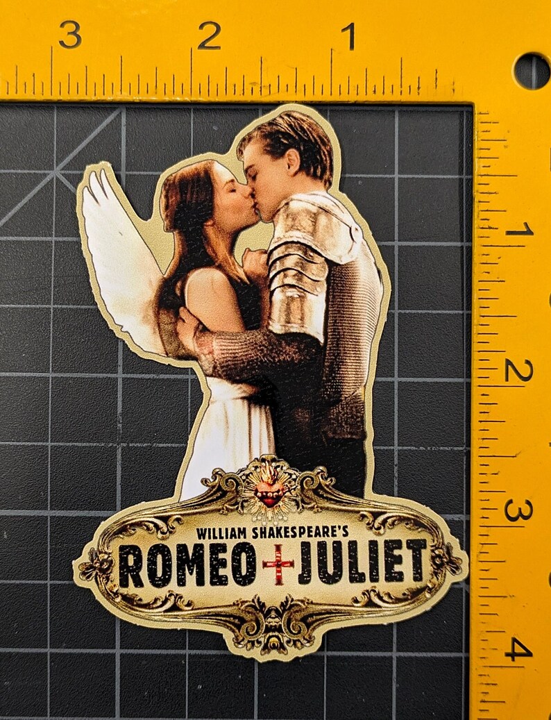 May include: A gold-coloured sticker with a decorative frame and the text "WILLIAM SHAKESPEARE'S ROMEO + JULIET". The sticker features a couple kissing, with a white angel wing behind them.
