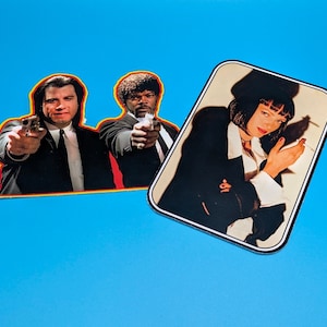 May include: Two stickers featuring characters from a film. One sticker shows two men in black suits pointing guns. The other sticker depicts a woman in a black suit holding a cigarette. The stickers have a white border and are set against a blue background.