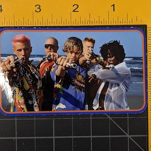 May include: A sticker featuring a group of four people on a beach, all wearing colourful shirts and holding toy guns. The people are smiling and looking at the camera. The background is a blue sky and ocean.