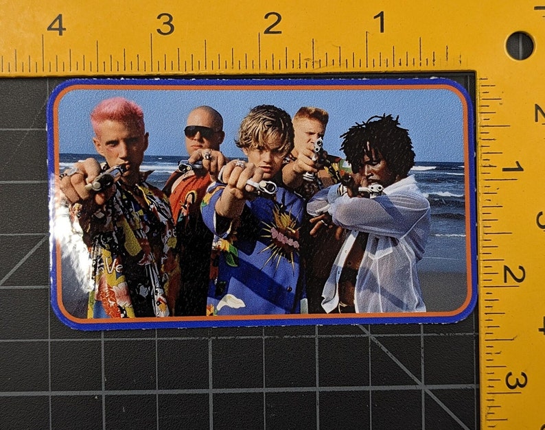 May include: A sticker featuring a group of four people on a beach, all wearing colourful shirts and holding toy guns. The people are smiling and looking at the camera. The background is a blue sky and ocean.