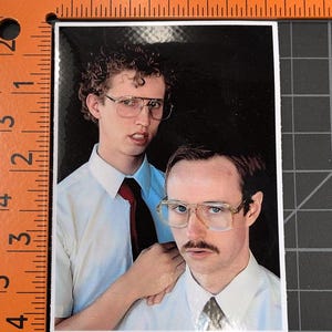 May include: Two men wearing white shirts and ties, one with curly brown hair and glasses, the other with brown hair and a mustache, are standing together. The man with curly hair is holding his hand on the other man's shoulder.
