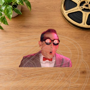 May include: A die-cut sticker featuring a person wearing black-rimmed glasses with red and white spiral lenses labeled X-RAY SPEX, a white collared shirt, a red bow tie, and a grey blazer, set against a light-toned wood grain background.