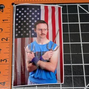 May include: A man with glasses and a mustache is holding two silver sai weapons in front of an American flag background. He is wearing a blue tank top.