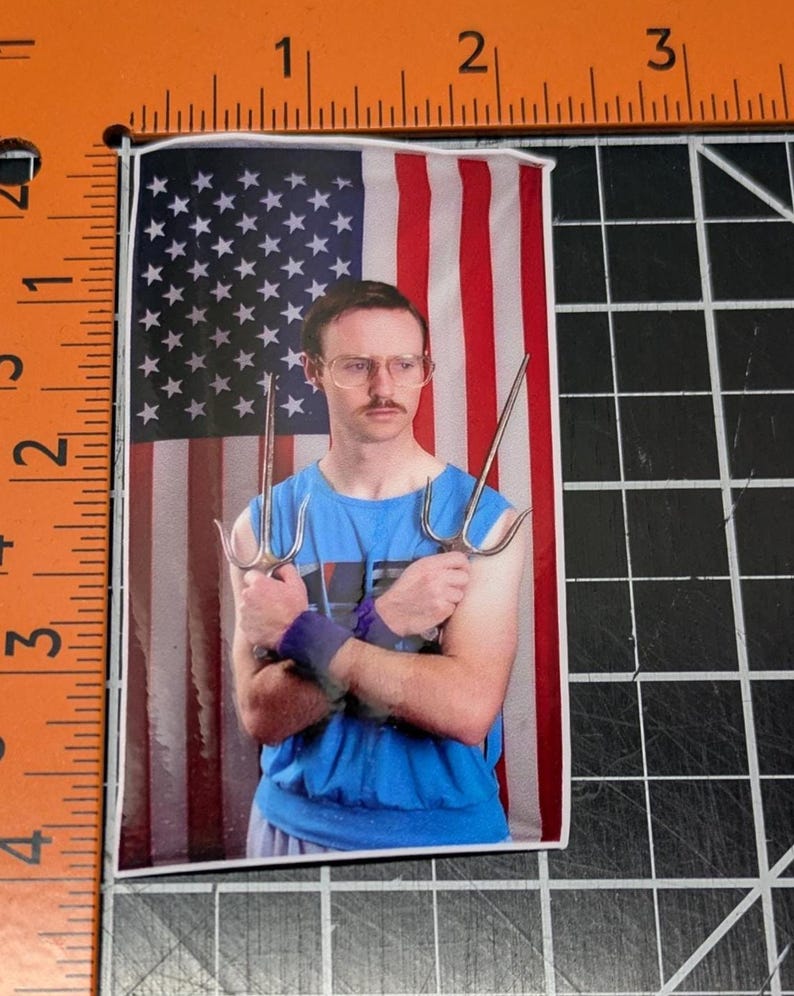 May include: A man with glasses and a mustache is holding two silver sai weapons in front of an American flag background. He is wearing a blue tank top.