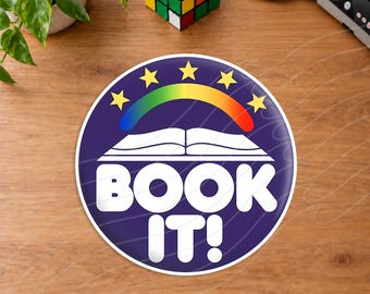 May include: A circular sticker with a dark blue background featuring a white open book graphic, a colorful rainbow arch, and five yellow stars above it. The text BOOK IT! is printed in large, bold white capital letters at the bottom of the sticker.
