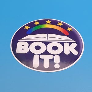 May include: A round sticker with a blue background and a white border. The sticker features the words "BOOK IT!" in white, with an open book design above the text. A rainbow arch and yellow stars complete the design.
