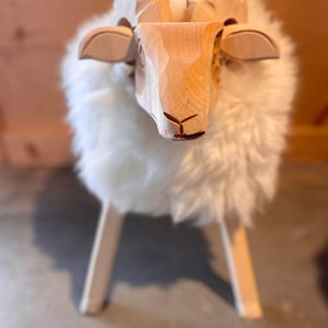 May include: A wooden sheep-shaped stool with a carved head and horns. The sheep's body is covered in fluffy white material. The stool has four pale wooden legs, and is a decorative piece.