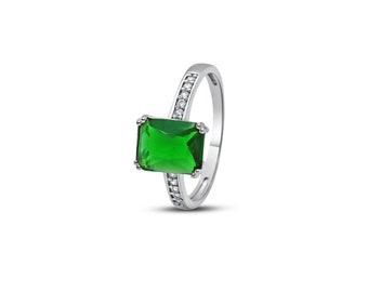 Puede incluir: Un anillo plateado con una gran gema rectangular de color verde esmeralda. La banda está adornada con pequeñas piedras claras. El anillo se presenta sobre un fondo blanco, resaltando su diseño.