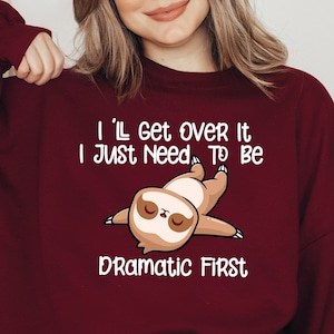 May include: Burgundy sweatshirt with the text "I'll get over it, I just need to be dramatic first" and a cartoon sloth illustration. The sloth is light brown and cream-colored. The sweatshirt is a long-sleeved crew neck.