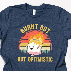 May include: A navy blue t-shirt with a vintage-style graphic of a burnt marshmallow with the text "Burnt Out But Optimistic".
