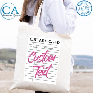 May include: A white tote bag featuring a library card design with the words "Custom Text" in pink script. The top of the card reads "Library Card" with spaces for "Title", "Date Due", and "Borrower Name". The bag has a "Conquerer Apparel" logo.