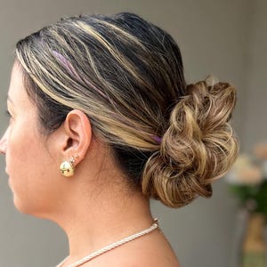Puede incluir: Una camiseta de tirantes negra con tirantes finos, un pendiente esférico dorado y un collar de cuentas delicado. El cabello está peinado en un moño bajo texturizado y voluminoso con reflejos rubios, y un tatuaje de rayos de sol es visible.