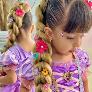 May include: A young girl with long, braided blonde hair adorned with colorful flower hair clips. She wears a purple princess dress with a gold trim and a cameo. The image is a close-up, showcasing the hairstyle and dress.