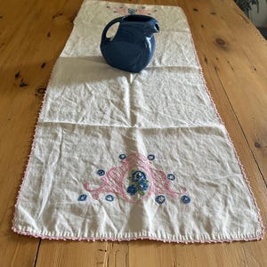 Puede incluir: Un camino de mesa rectangular de tela blanca con bordados florales en rosa y azul en ambos extremos y un delicado borde de ganchillo rosa. Una jarra de cerámica azul oscuro con asa curva y pico vertedor se sitúa sobre el camino.