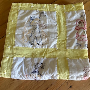 May include: A handmade, quilted baby blanket with embroidered nursery rhyme characters. The blanket features yellow borders and white squares with embroidered figures of "Boy Blue" and "Simple Simon". The blanket is on a wooden surface.