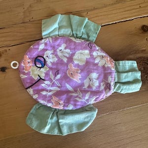 May include: A handmade fish-shaped item with a lavender floral pattern body and light green ruffled fins. The fish has a white eye, a blue embroidered eye, and a black embroidered smile. The item is likely a decorative piece or a toy.
