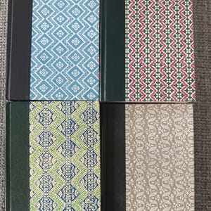 May include: Four hardback books with patterned covers in blue, red, green, and beige. The spines are solid colours, including black and green. The books are arranged in a square formation, showcasing their decorative designs.
