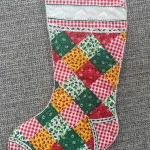 Puede incluir: Un calcetín navideño acolchado con un diseño de patchwork de cuadros vichy rojos y blancos, estampado floral amarillo y hojas de acebo verdes. El puño superior es blanco con un borde de cuadros vichy y una textura de rombos acolchados.