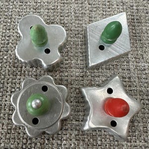 Può includere: Quattro formine per biscotti vintage in metallo di varie forme: un fiore, un diamante, un cerchio smerlato e una stella. Ogni formina ha un pomello in legno colorato. Le formine a fiore e a diamante hanno pomelli verdi, la stella un pomello rosso.