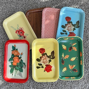 Puede incluir: Una colección de bandejas metálicas vintage en varios colores y diseños. Incluye una bandeja roja con rosas naranjas, una bandeja amarilla con rosas rojas, una bandeja azul con rosas, una bandeja verde con hojas de otoño, una bandeja marrón y una bandeja a cuadros rojos y blancos.